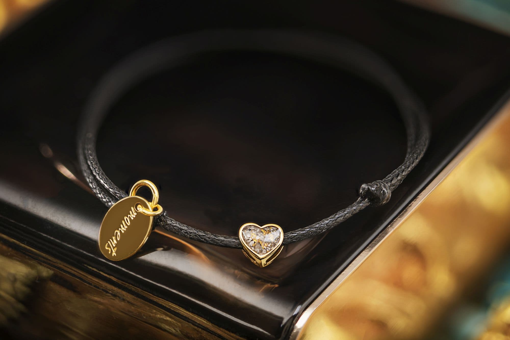 Geflochtenes schwarzes Kordelarmband mit goldenem ovalen Anhänger und goldenem Herzanhänger, der Asche enthält.