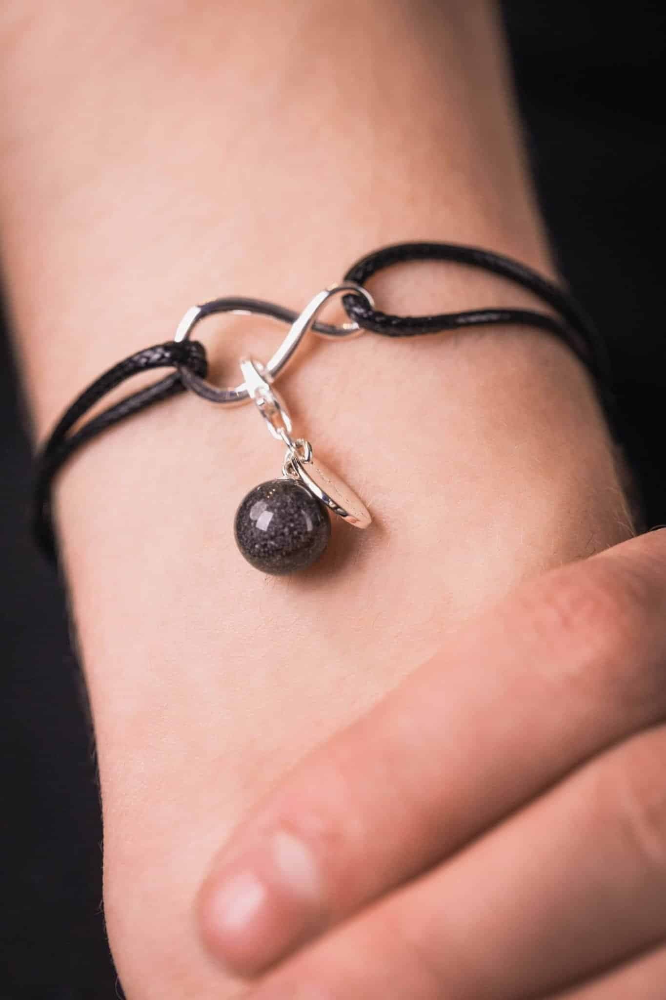Schwarzes Kordelarmband mit silbernem Unendlichkeitssymbol, mit einer gesprenkelten dunkelgrauen Gedenkperle und einem kleinen silbernen Scheibenanhänger.