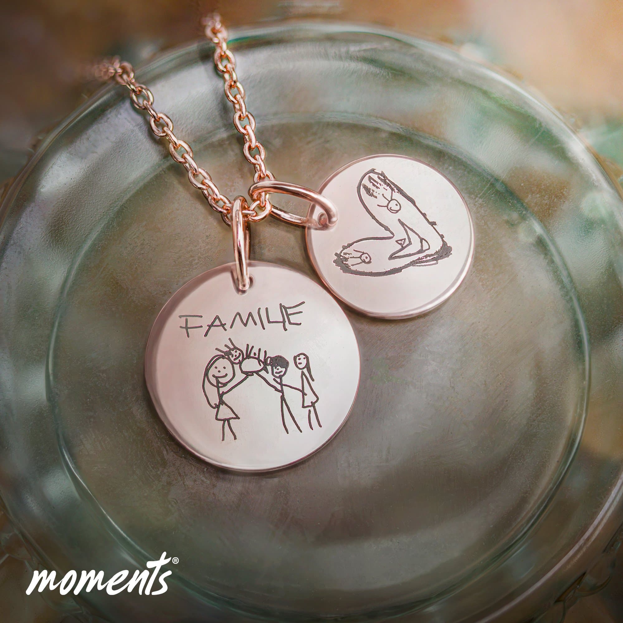 Roségoldene Halskette mit zwei runden Anhängern, einer graviert mit 'FAMILE' und einer Strichmännchen-Familie, der andere mit einer Kinderzeichnung.