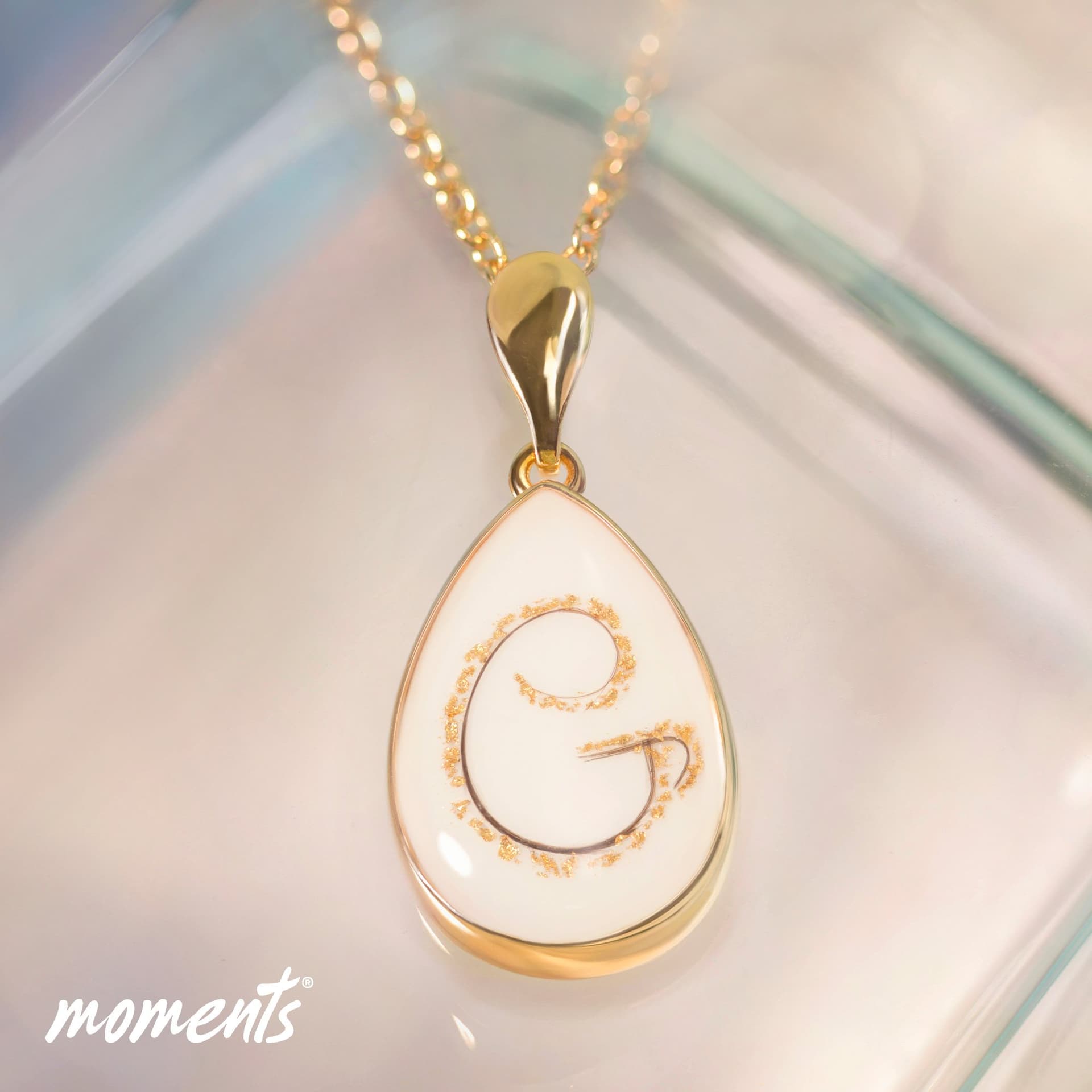 Gouden druppelvormige hanger met witte inleg, met een donkere "G" initiaal en gouden spikkels.