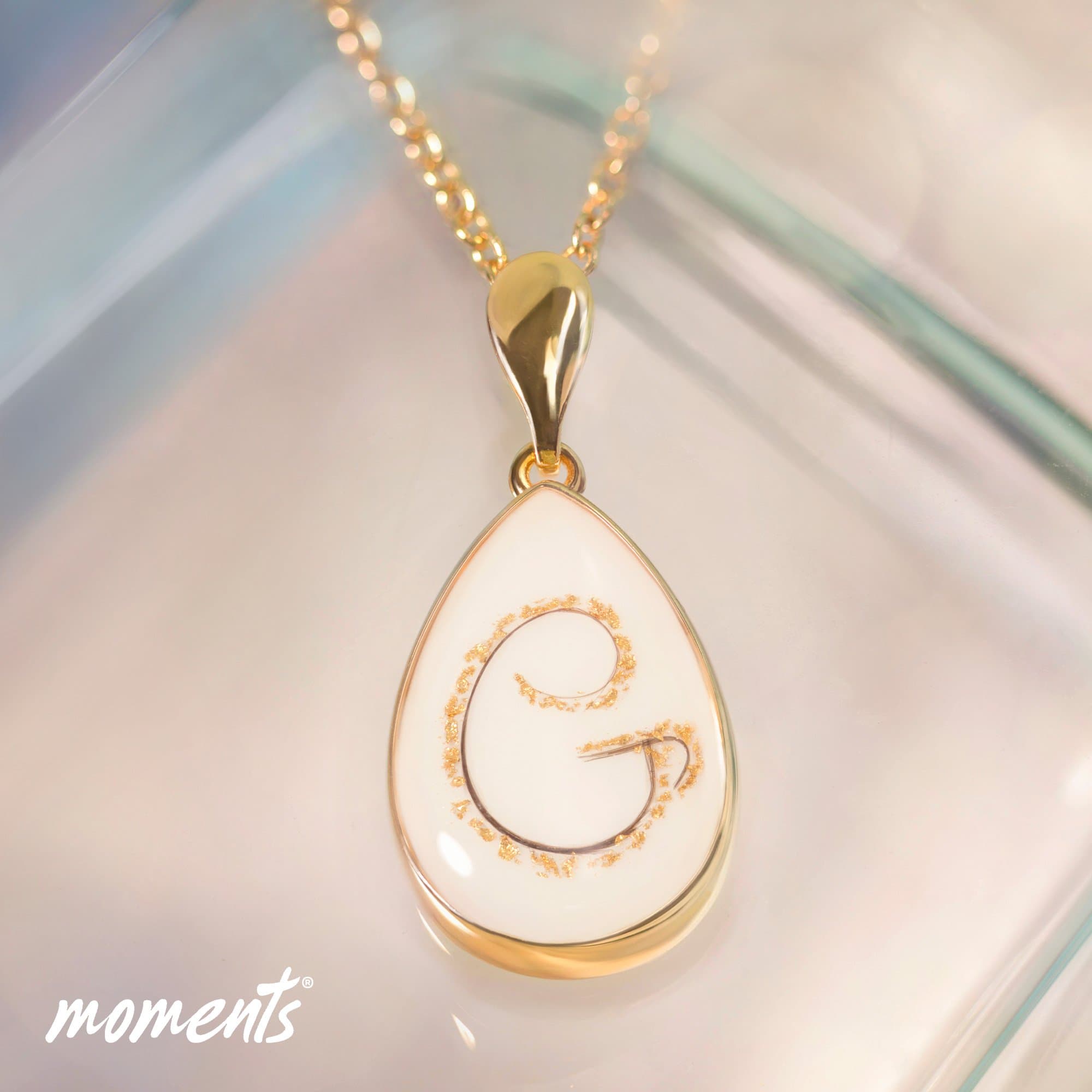 Goldener tropfenförmiger Anhänger mit weißer Einlage, mit dunkler "G" Initiale und Goldsprenkeln.