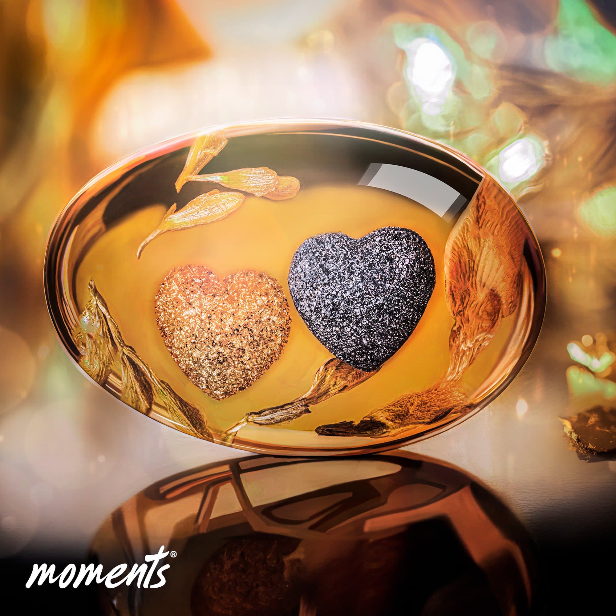 Ovales Bernsteinharz mit zwei Glitzerherzen, eines in Gold, eines in Schwarz, und getrockneten Pflanzenelementen.
