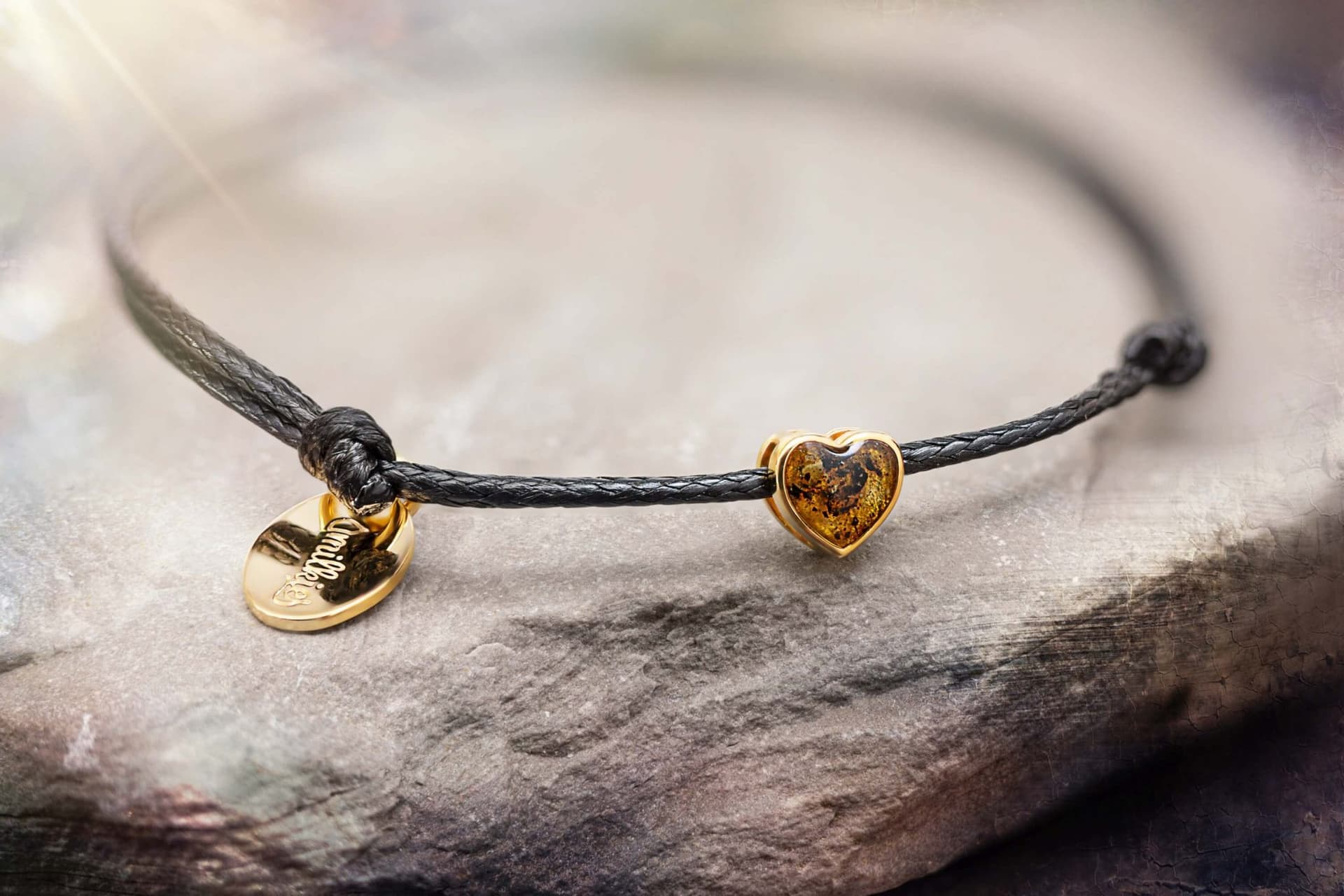Armbånd med sort bånd, en gylden hjerteformet rav-charm og et ovalt gyldent vedhæng.