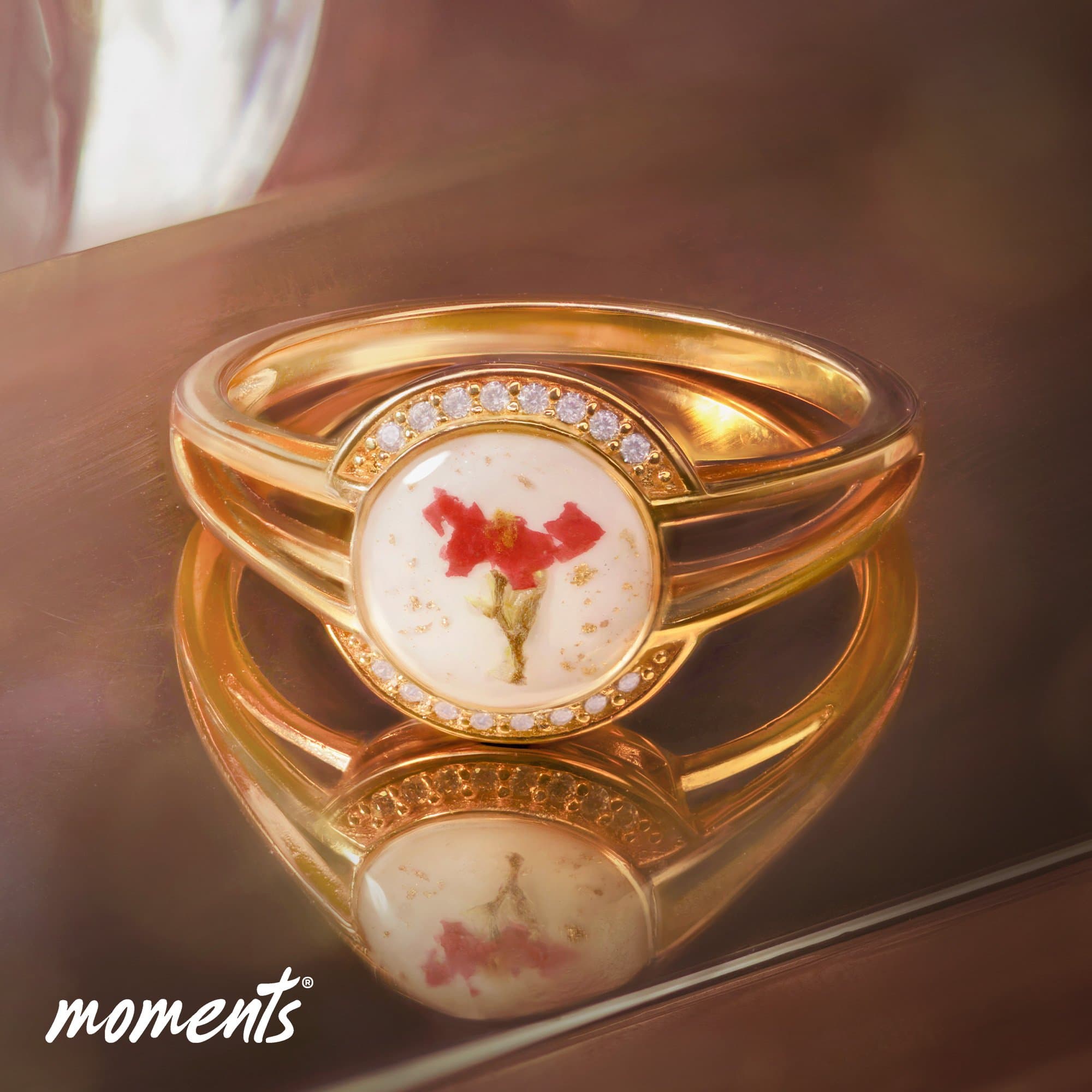 Goldring mit weißem rundem Element, das eine rote Blüte und Goldflecken enthält, umgeben von kleinen klaren Steinen.