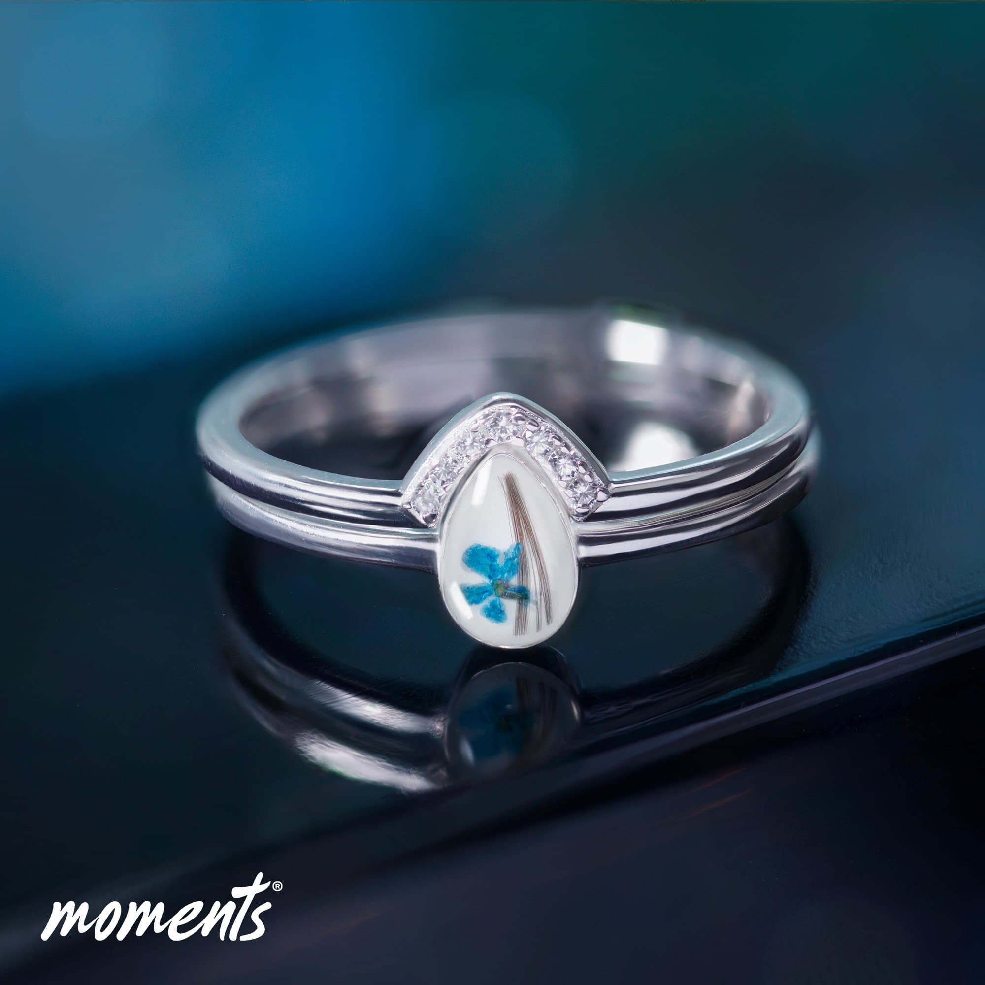 Silberring mit tropfenförmigem klarem Stein, der eine blaue Blüte und Haarsträhnen enthält.