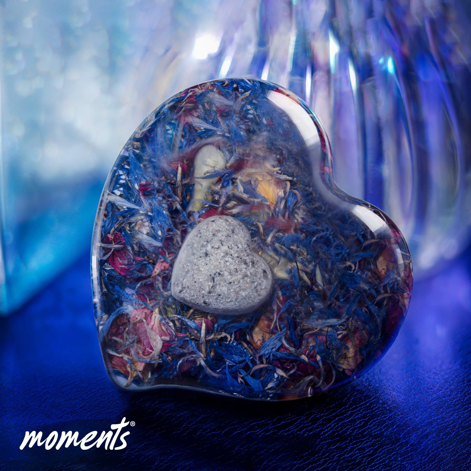 Herzförmiges Erinnerungsstück aus Harz mit blauen und rosa Blütenblättern und einem kleinen grauen Herzstein.