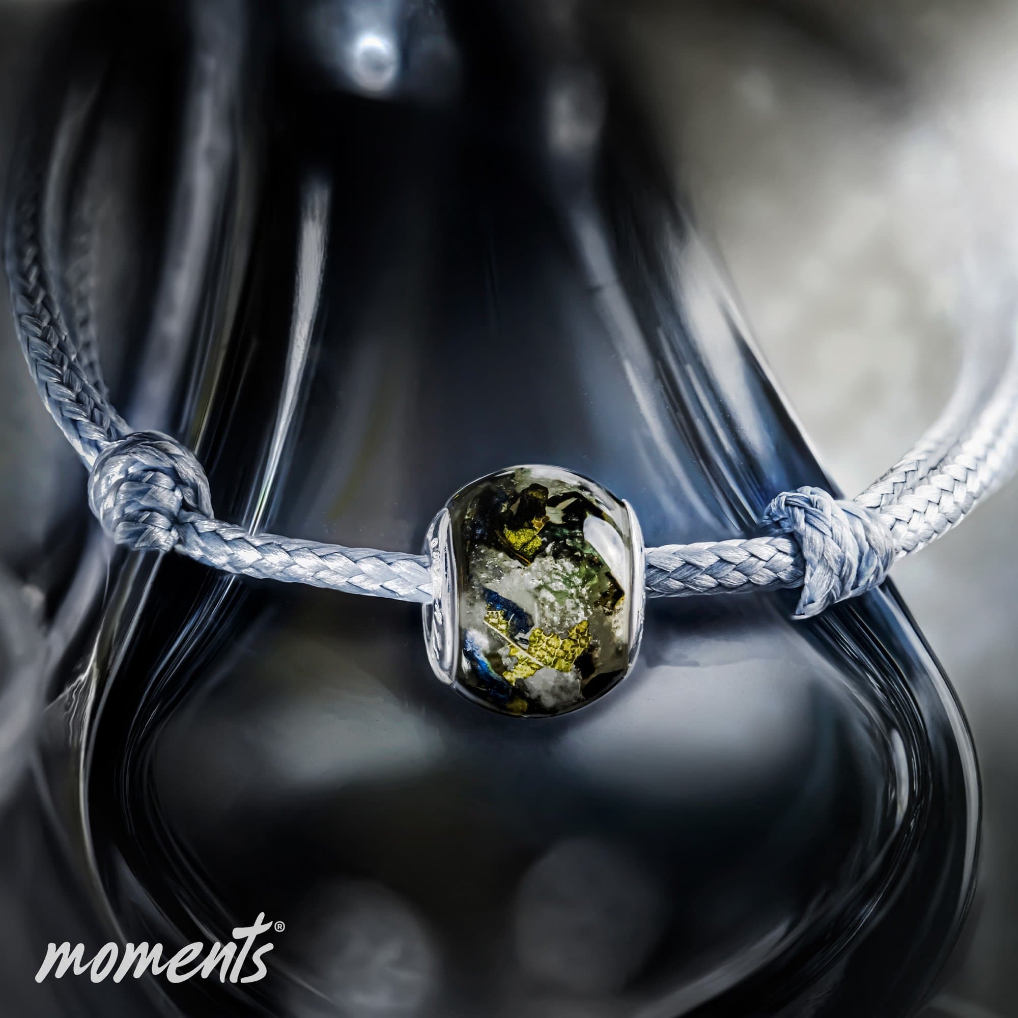 Glasperle mit eingeschlossenen grünen und goldenen Elementen an einem geflochtenen silbernen Band.