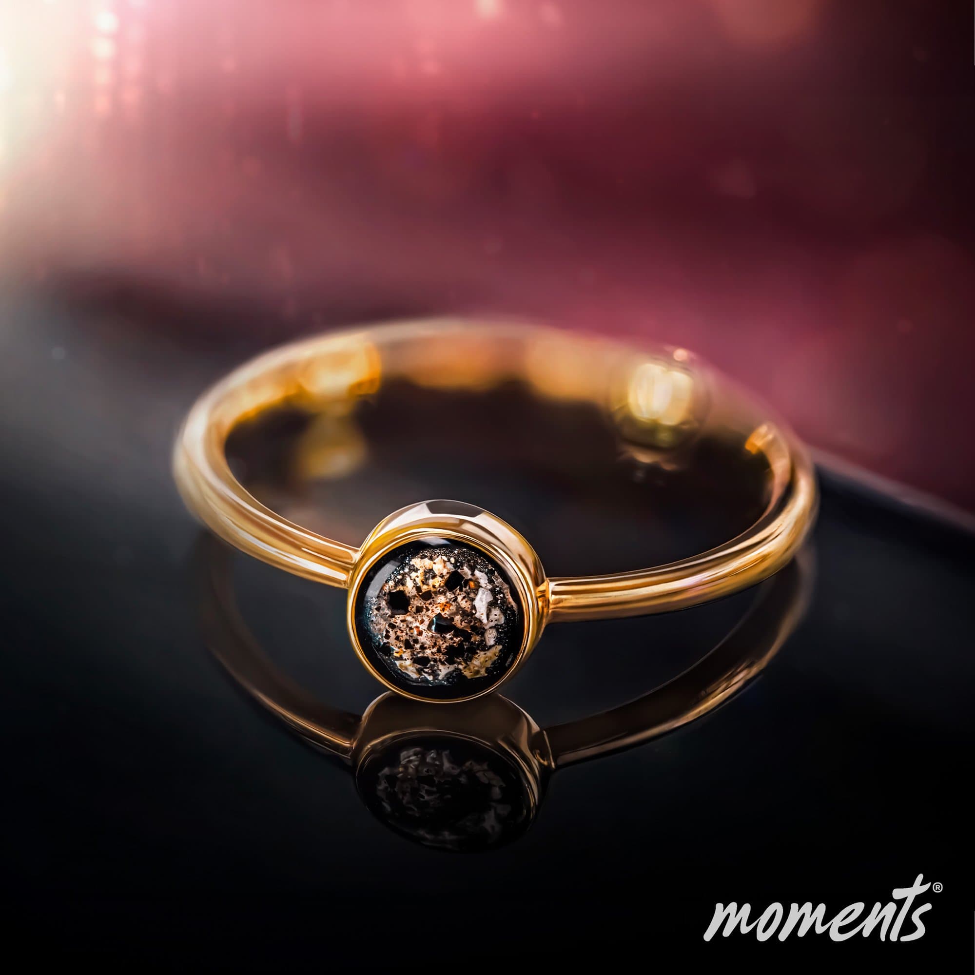 Goldring mit einem runden, dunklen Erinnerungsstein mit gold- und weißfarbenen Flecken.