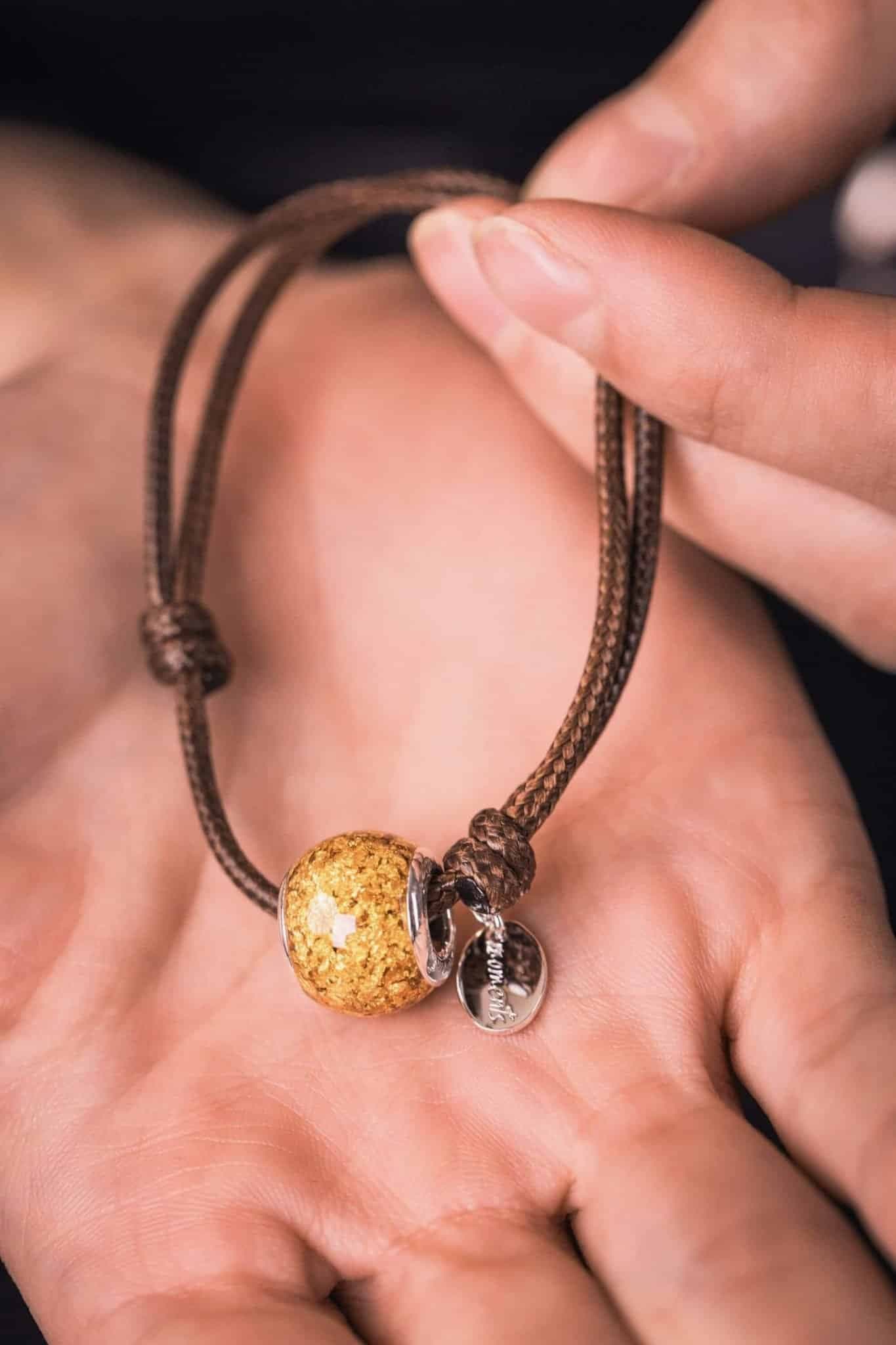 Braunes Kordelarmband mit einer goldenen, strukturierten Perle und einem silbernen Anhänger.