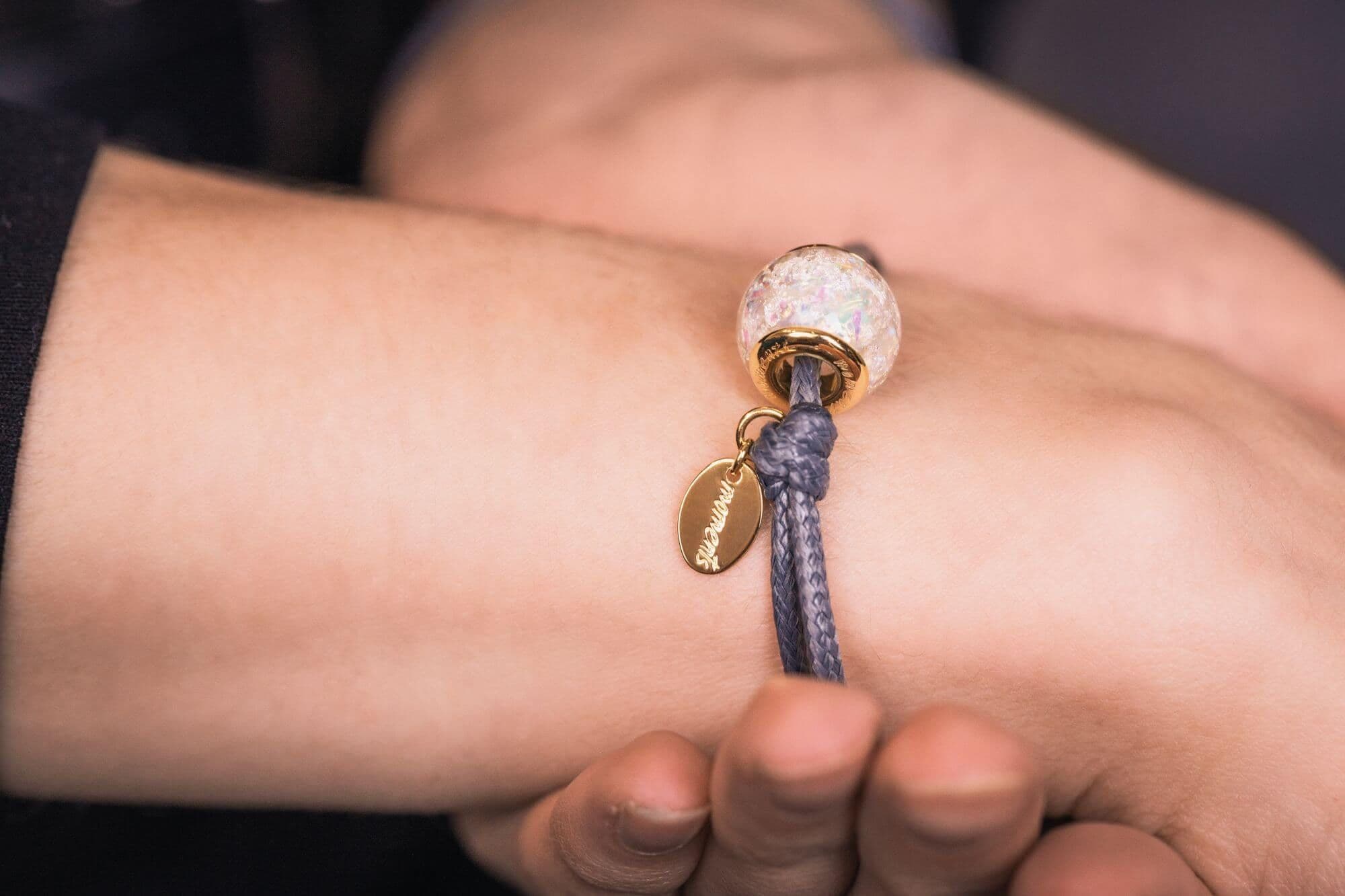 Glasperle mit irisierenden Einschlüssen an einem grauen Kordelarmband mit einem goldenen ovalen Anhänger.