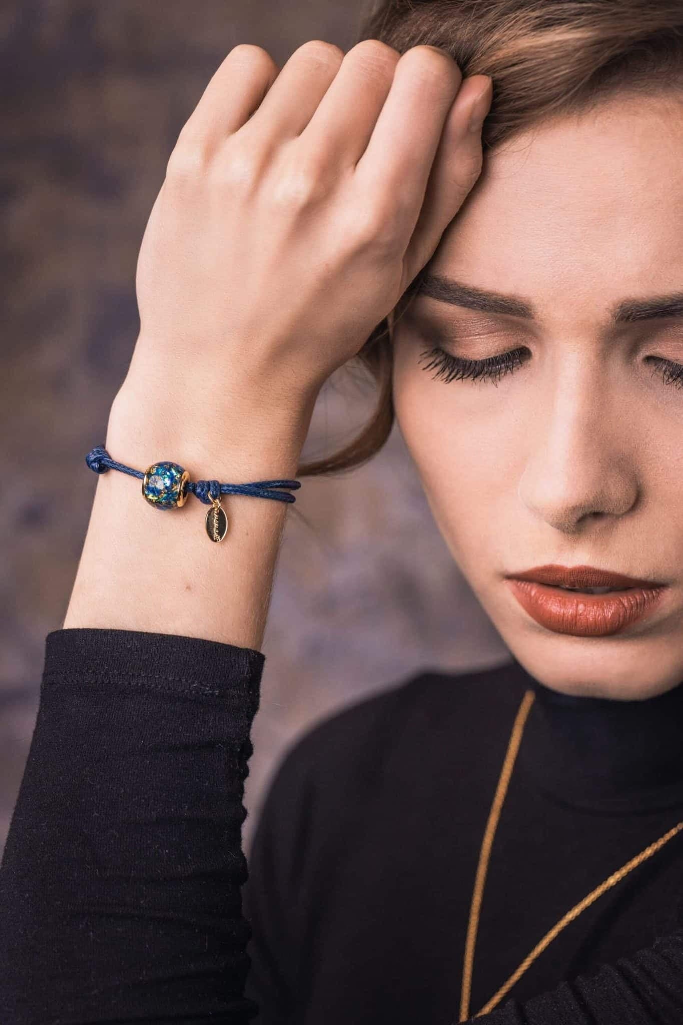 Blaues Kordelarmband mit einer goldfarbenen Perle, die blaue und goldene Flocken enthält, und einem kleinen ovalen Anhänger.