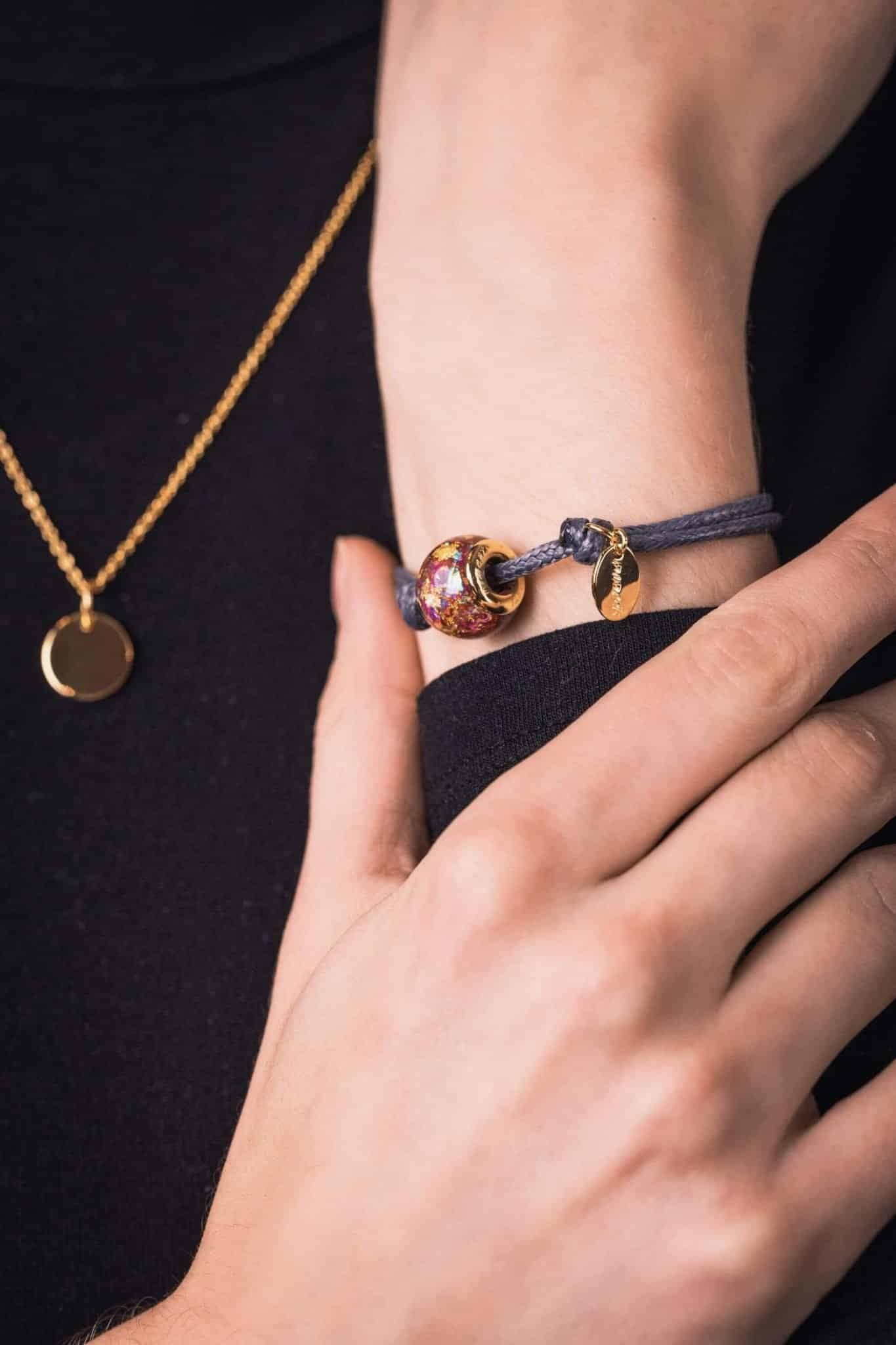 Eine vergoldete Perle mit roten und goldenen Flocken an einem grauen Kordelarmband.
