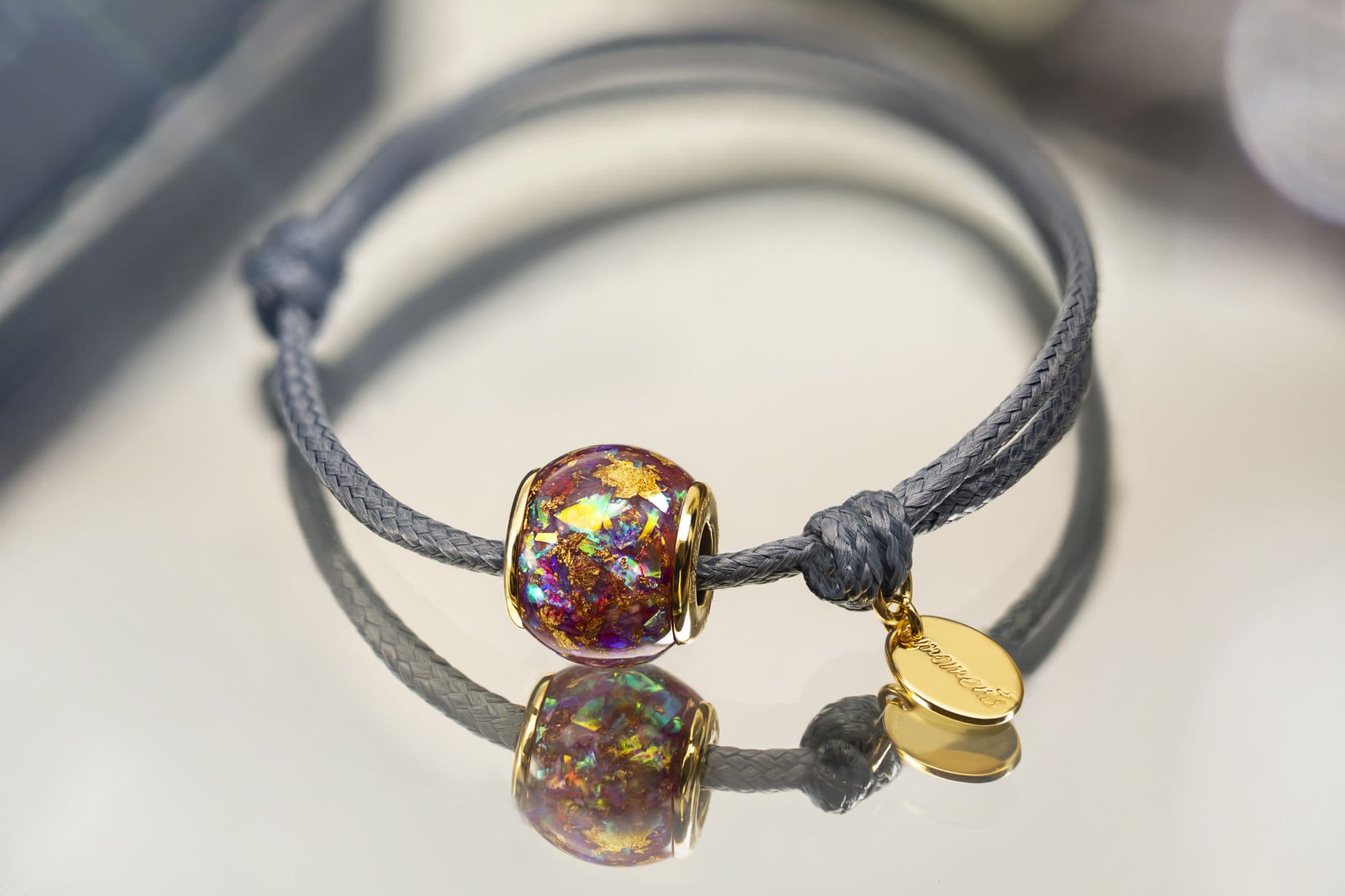 Graues Kordelarmband mit einer runden, mehrfarbigen Glasperle, die Goldflocken enthält, und einem goldenen ovalen Anhänger.