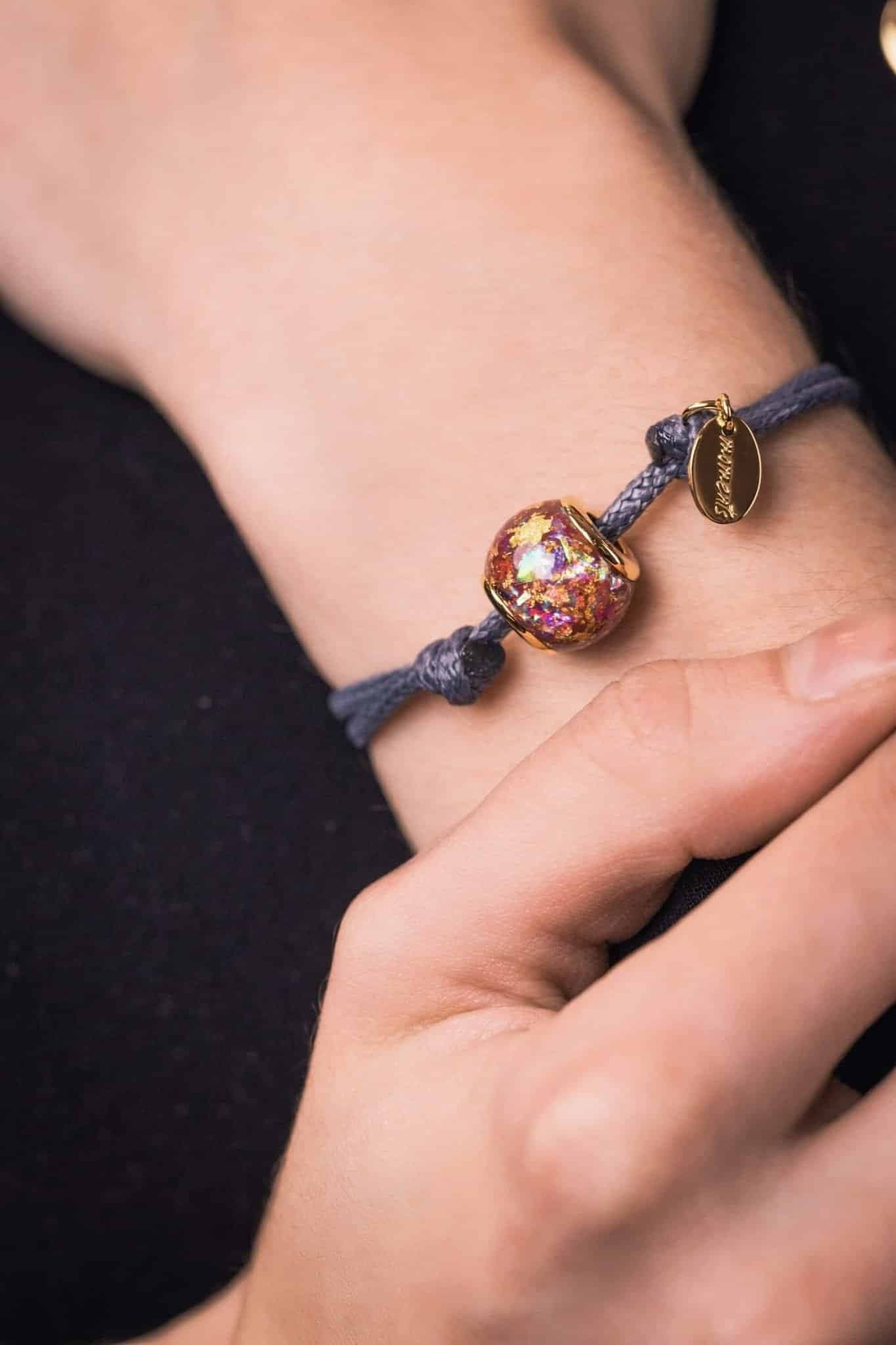 Graues Kordelarmband mit einer runden, irisierenden Perle und goldfarbenem Anhänger.