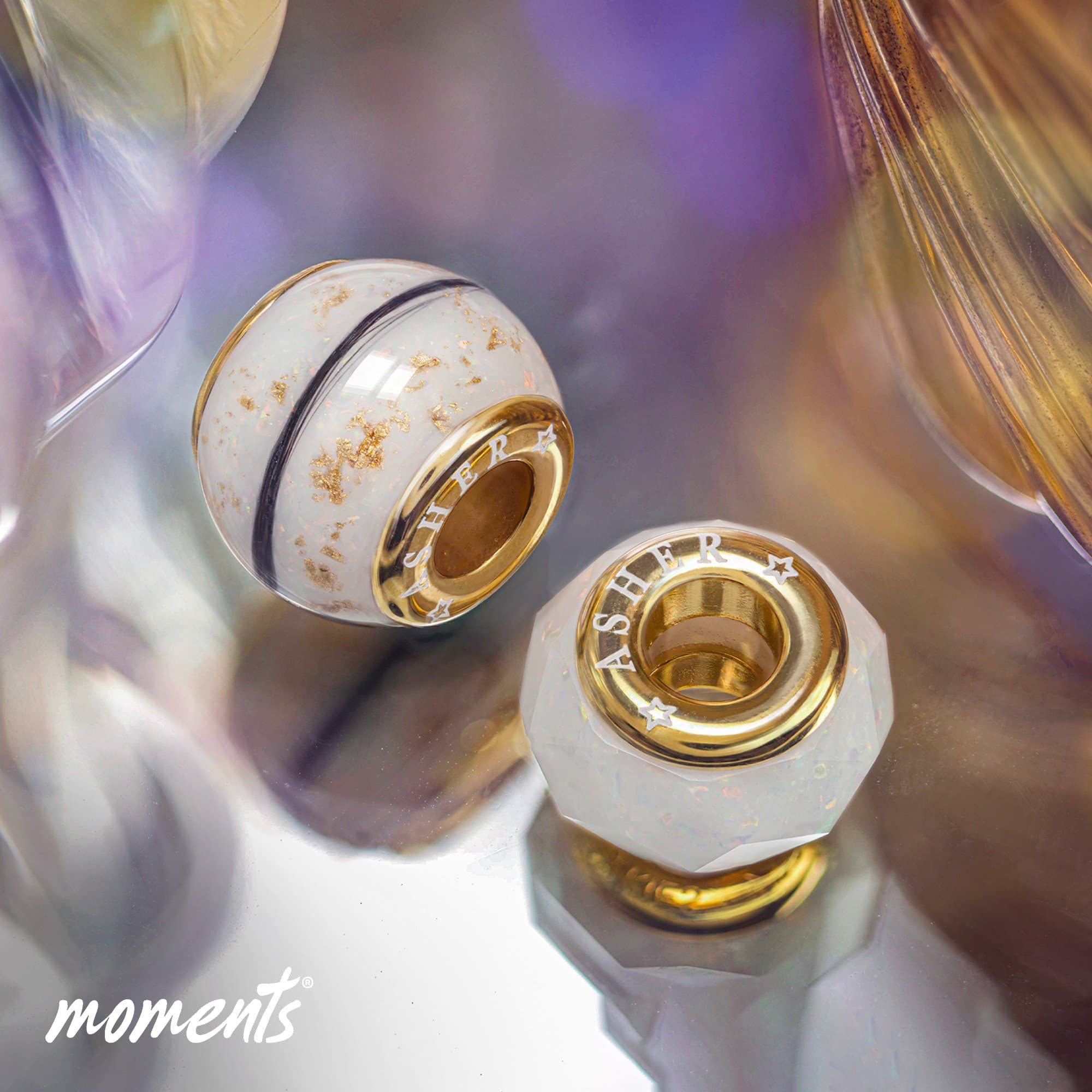 Zwei goldfarbene Anhänger, einer rund mit weißer Einlage und goldenen Flecken, einer facettiert mit weißer Einlage und Sternen.
