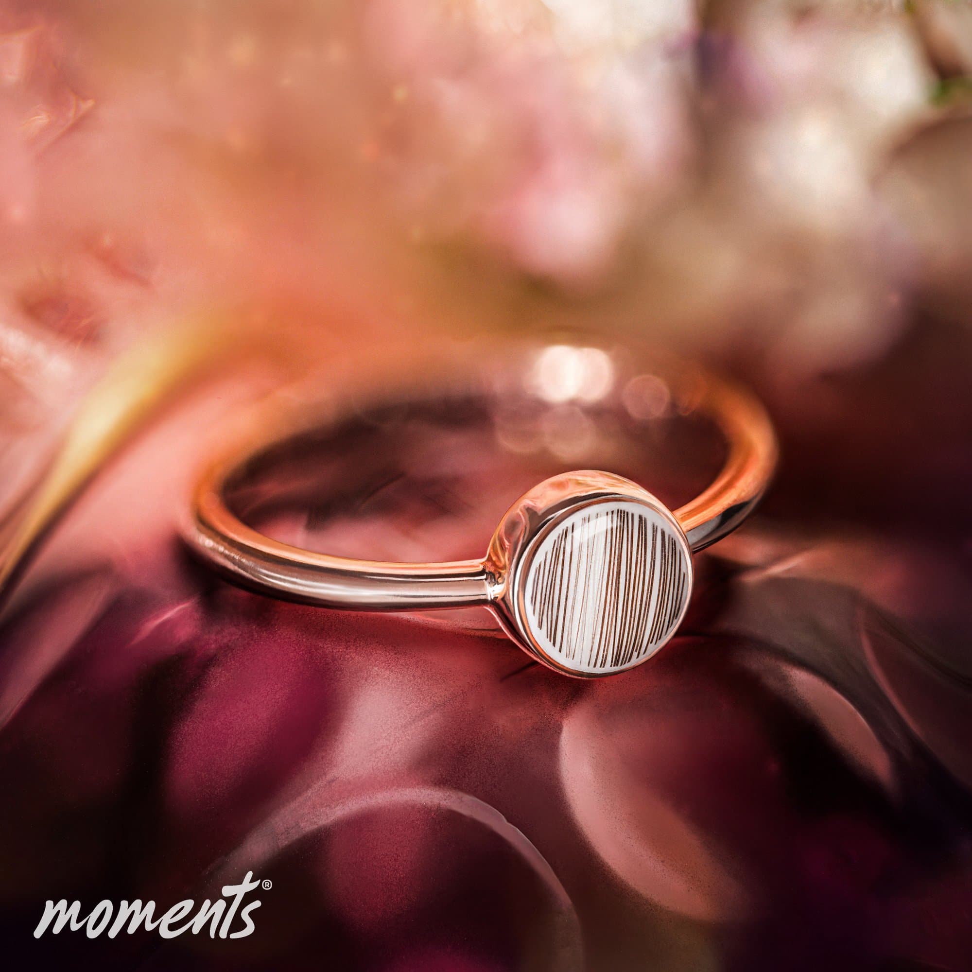 Roségoldener Ring mit einer runden Einlage aus weißem Material, das dünne, dunkle vertikale Linien aufweist.