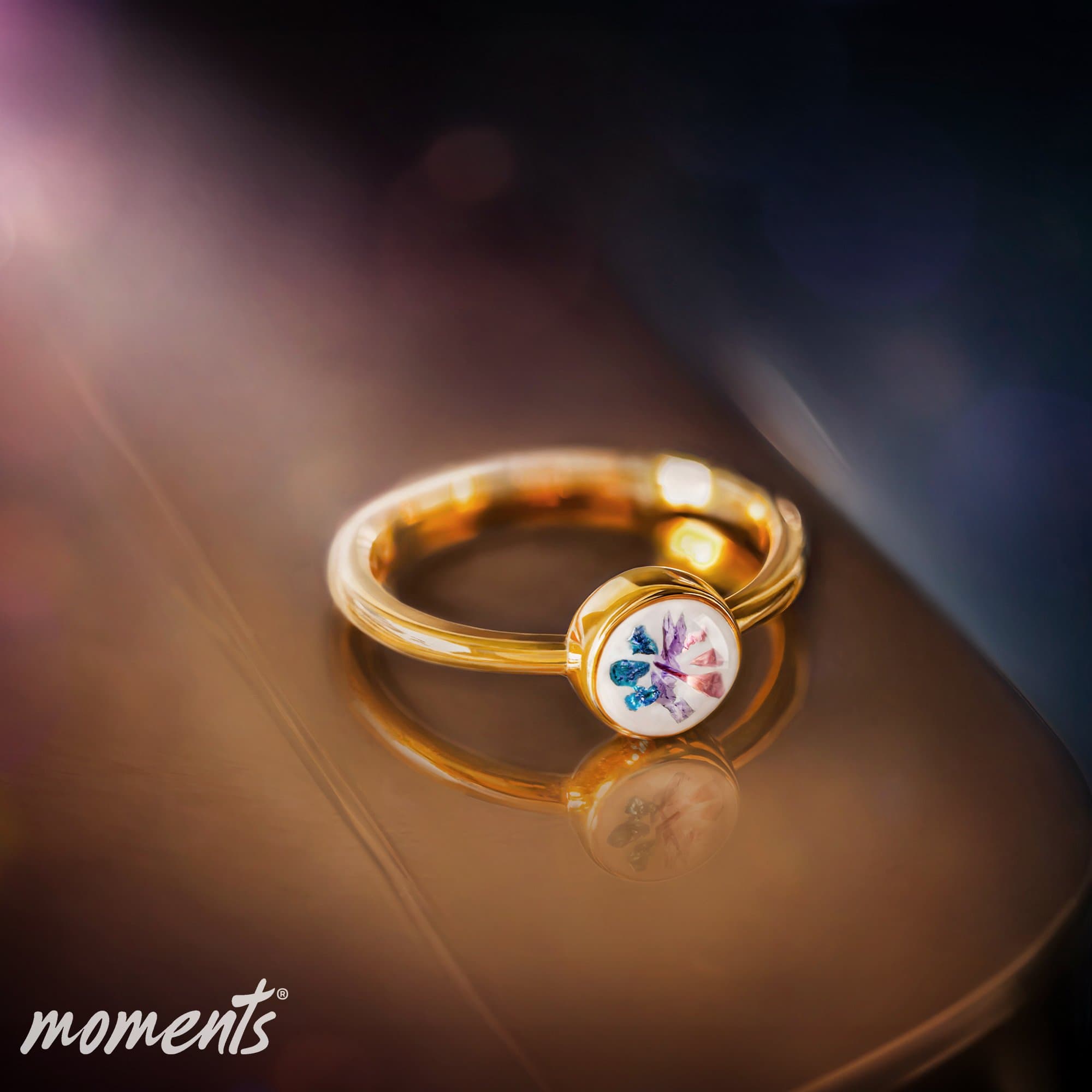 Goldring mit einer weißen runden Fassung, die bunte Fragmenten enthält.