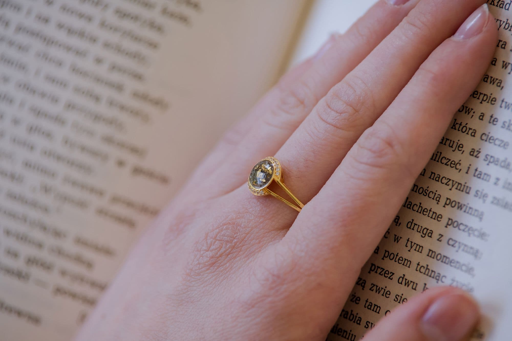 Goldring mit einem runden, grünen Erinnerungsstein mit goldenen Sprenkeln, umgeben von kleinen, klaren Steinen.