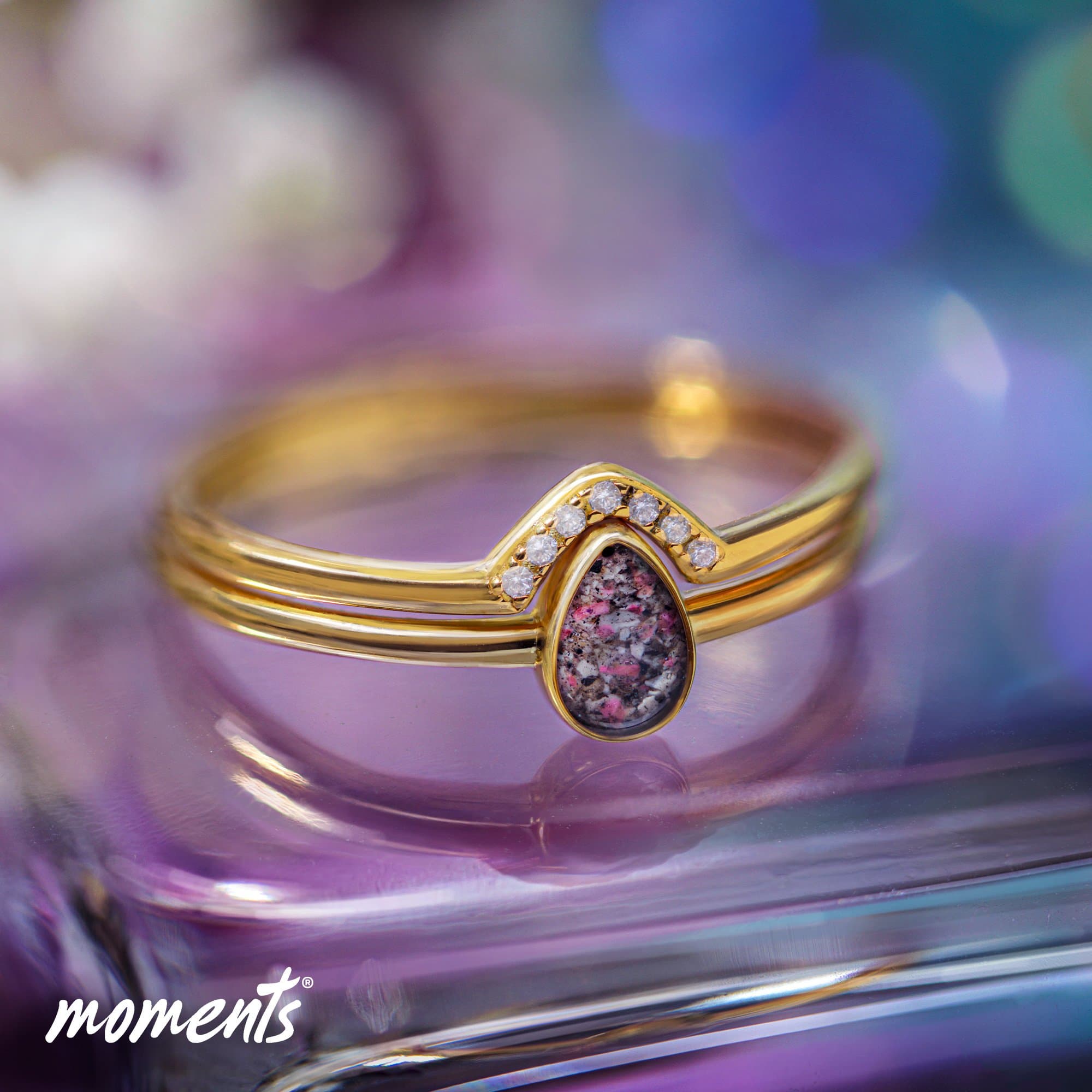 Goldring mit einem tropfenförmigen Erinnerungsstein mit rosa und grauen Sprenkeln, akzentuiert durch kleine, klare Steine.